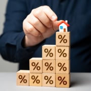Houten blokken met procenttekens opgestapeld als een trap, met een miniatuurhuisje bovenop, als symbool voor stijgende hypotheekrente