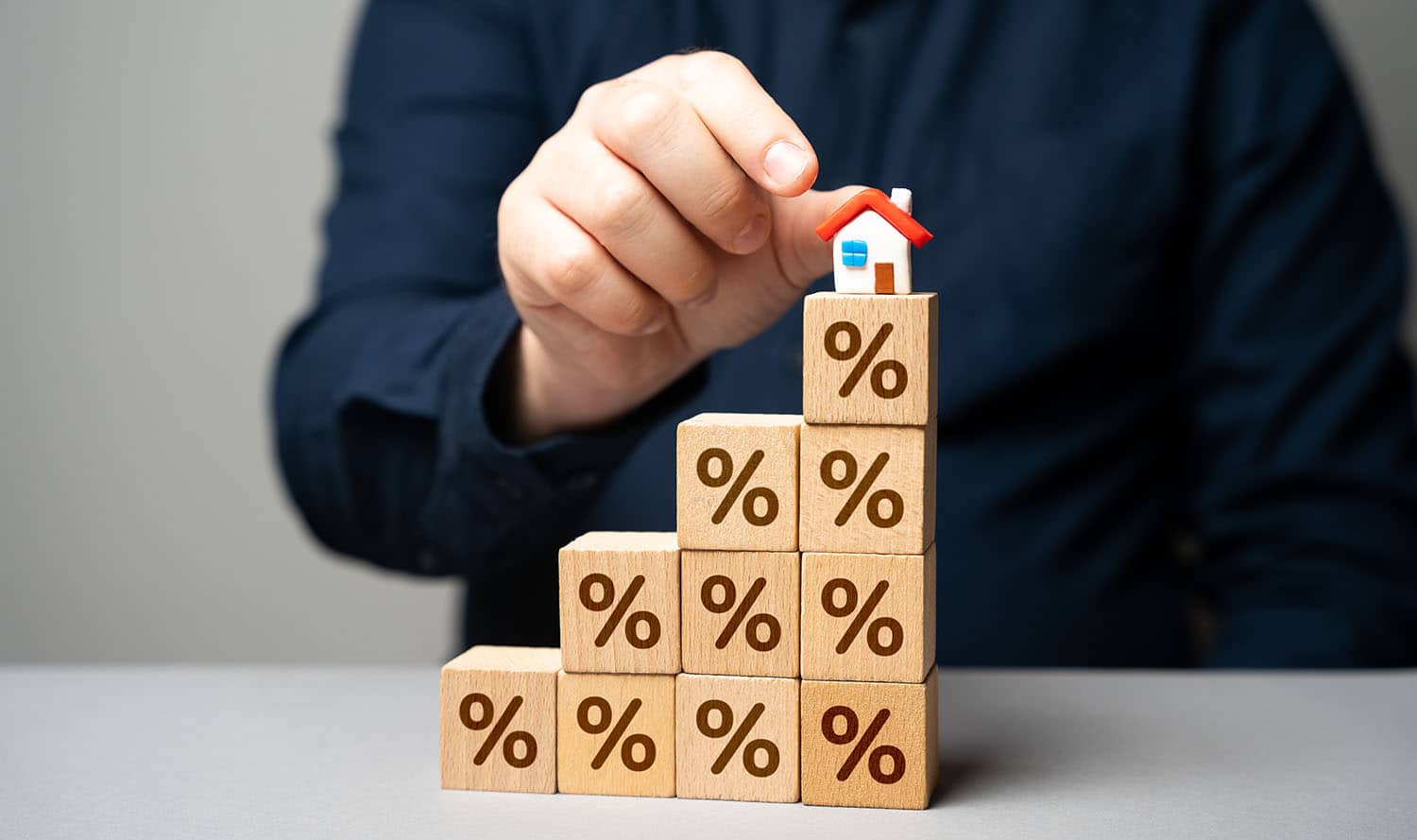 Houten blokken met procenttekens opgestapeld als een trap, met een miniatuurhuisje bovenop, als symbool voor stijgende hypotheekrente